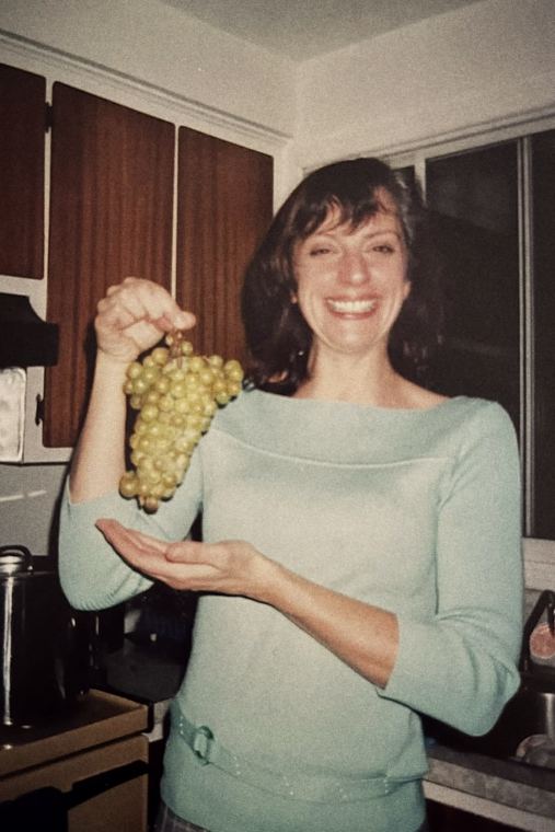 Josée en chandail vert d'eau, présentant une grappe de raisins verts et un rire.