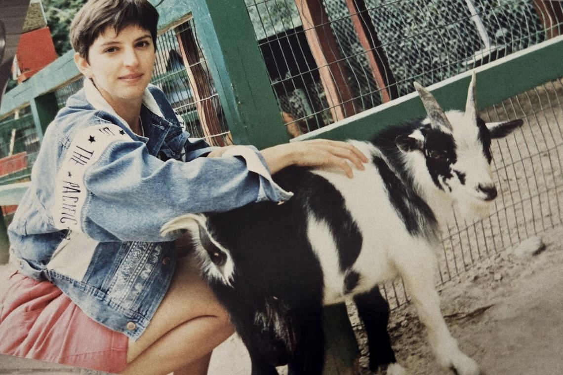 Josée en veste de jean, accroupie, la main sur le dos d'une chèvre noir et blanc.