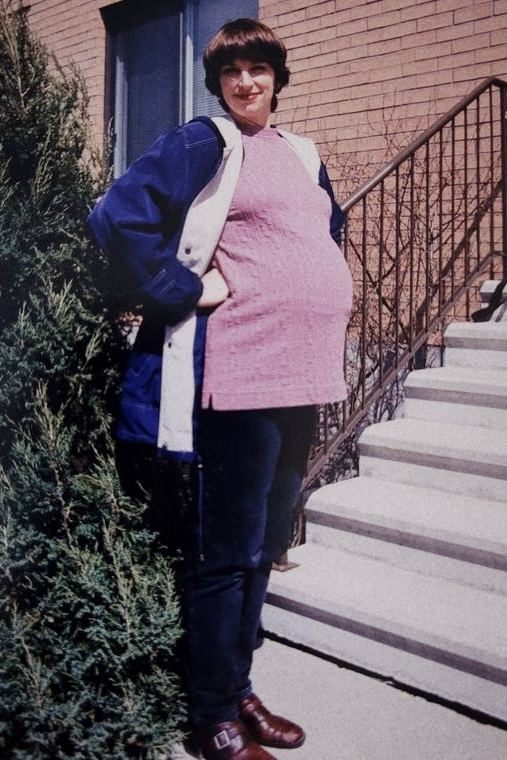 Josée enceinte, en tricot rose, devant un escalier de béton et un mur de briques.