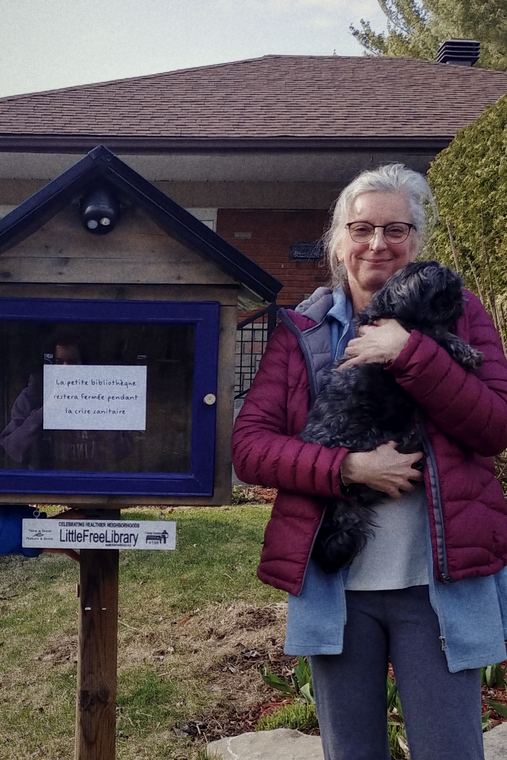 Josée devant une petite bibliothèque libre-service, son chien noir dans les bras.